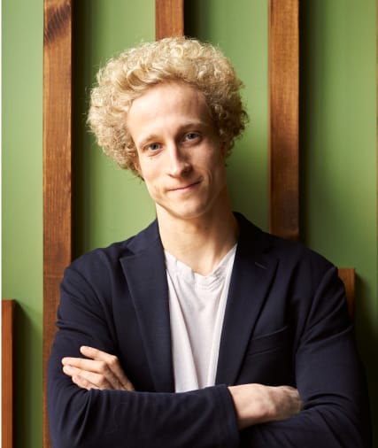 Young man with curly blond hair in a dark blazer crossing his arms, standing against a green background with wooden vertical panels.